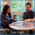 Two people playing a board game together with wine glasses, set against a windowed wall with greenery outside.